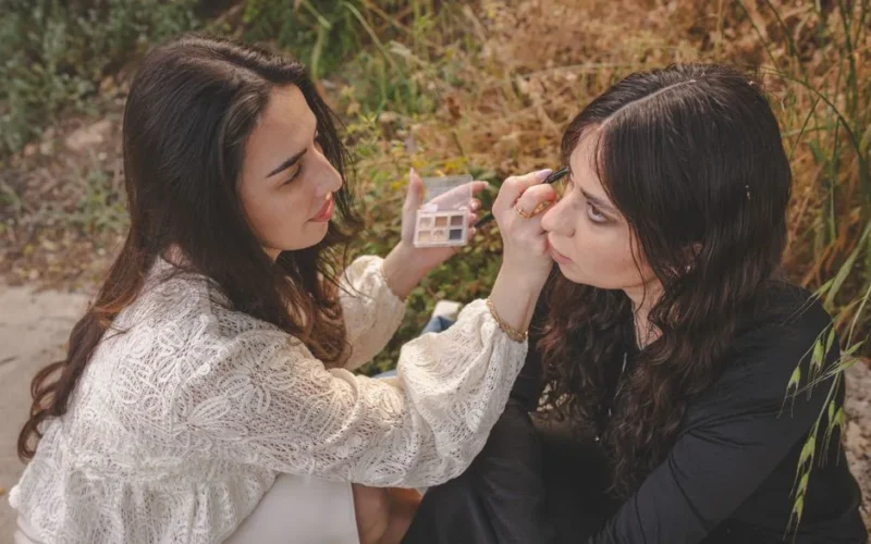 Two women outdoors, one applying makeup to the other, in a natural setting.