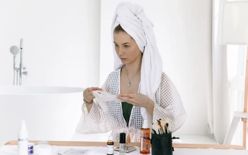 Woman with towel on head applying skincare products in a bright, minimalist bathroom setting.