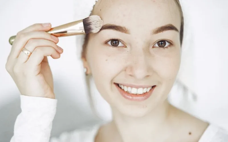 Portrait of a smiling young woman applying makeup with a brush indoors.
