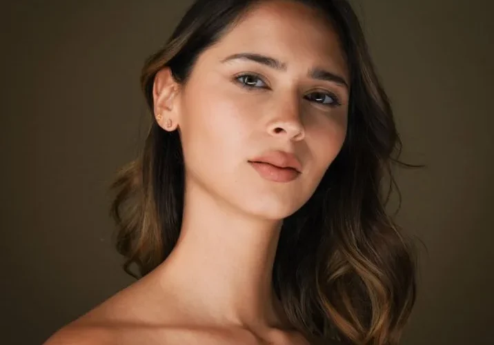 Studio portrait of a young woman with natural makeup, elegant pose, and soft lighting on a brown background.