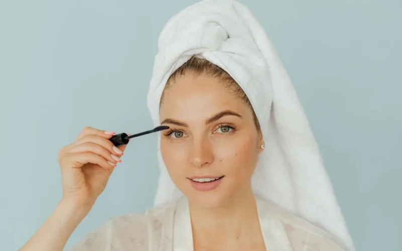 A woman smiles while applying mascara, wrapped in a towel headwrap against a light blue background.