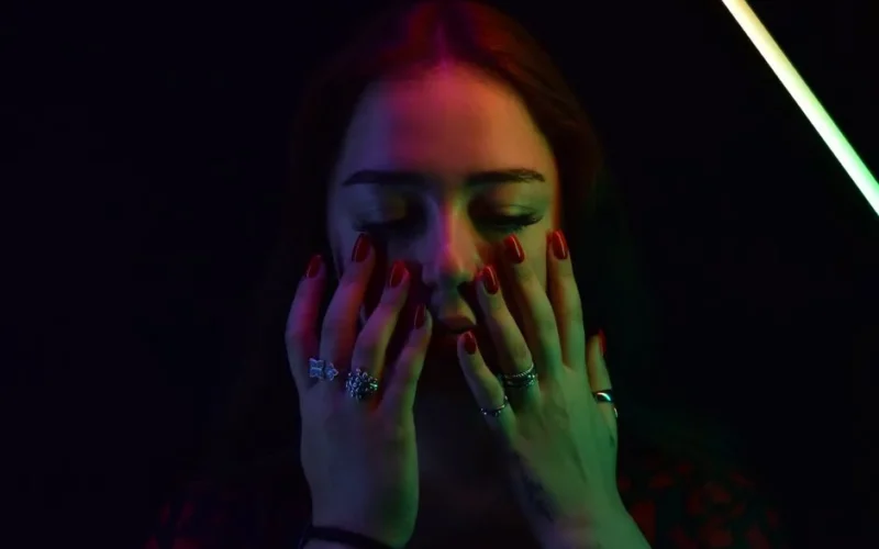Close-up portrait of a woman with colorful neon lights and closed eyes, showcasing rings and red nails.