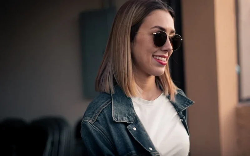 Fashionable young woman with short hair and sunglasses, smiling outdoors in a casual denim jacket.