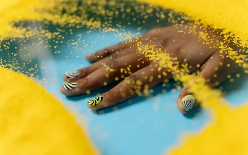 Artistic nail design with yellow and black patterns, surrounded by vibrant yellow sand on a blue surface.
