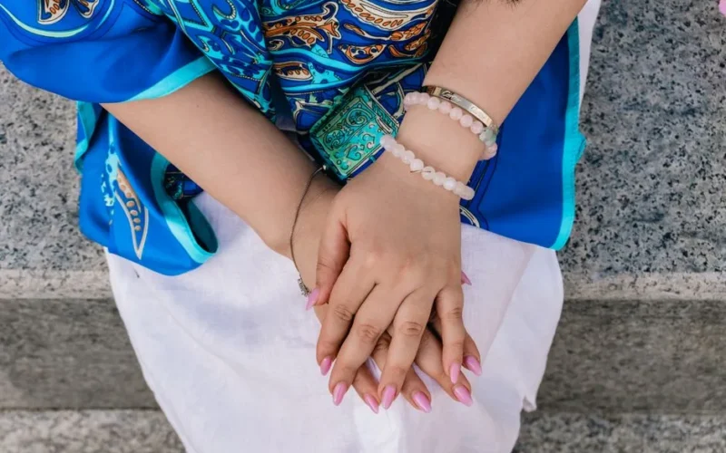 Close-up of a woman's hands with pink nails, wearing bracelets and a blue patterned dress.