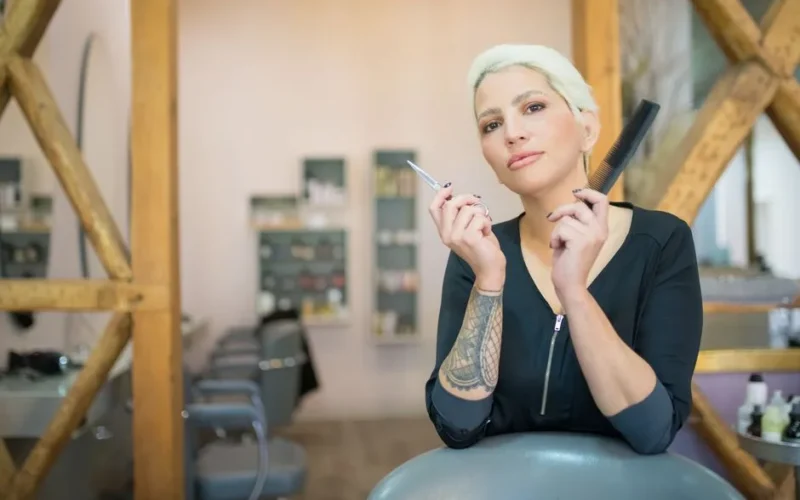 A confident hairdresser with arm tattoo and colored hair holding scissors and comb in a modern salon.