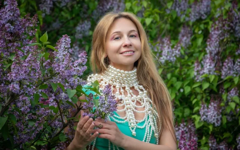 Elegant woman in pearl necklace posing with lilac flowers in a garden setting.