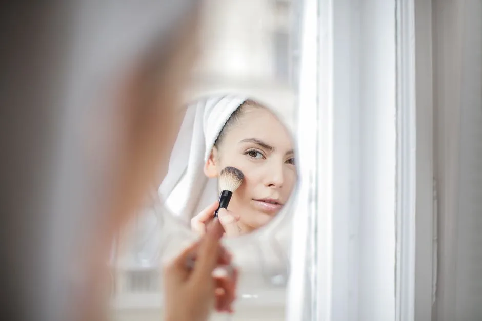 Reflection on mirror of young female using brush on face while applying makeup
