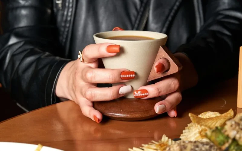 Close-up of manicured hands holding a coffee cup on a wooden table with snacks.