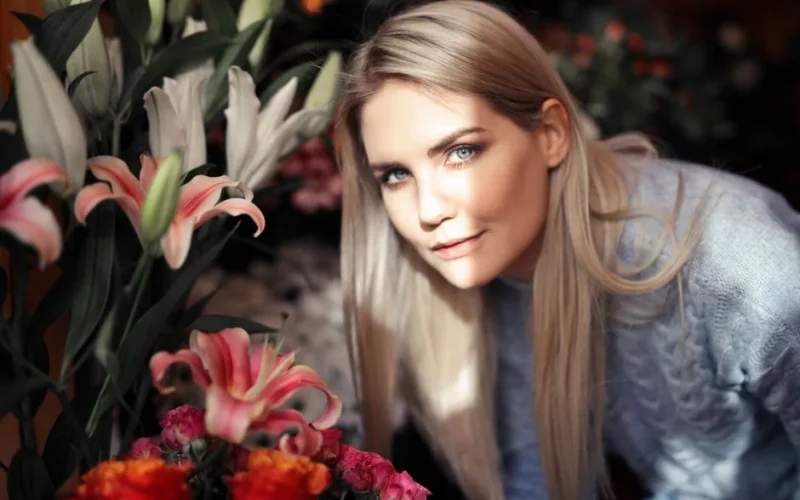 A blonde woman with blue eyes surrounded by a vibrant floral arrangement.