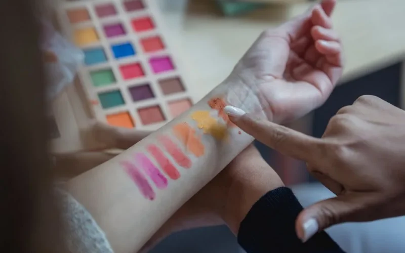 From above of crop unrecognizable young female makeup artist pointing at arm of client while applying colorful eyeshadows on wrist for testing