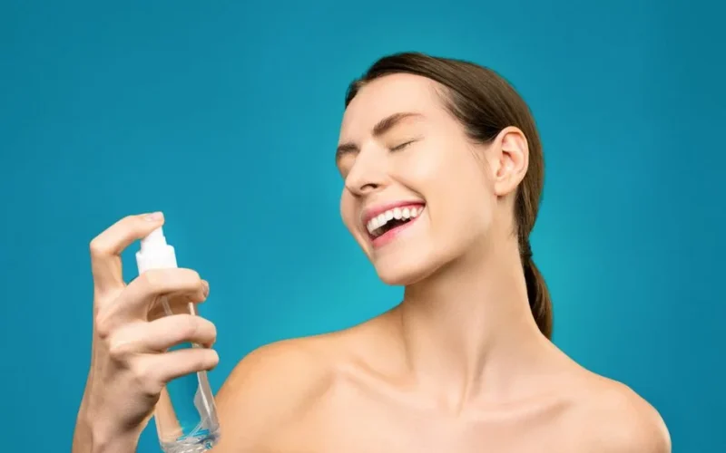 Close-up of a joyful woman spraying facial mist against a vibrant blue backdrop. Ideal for skincare promotions.