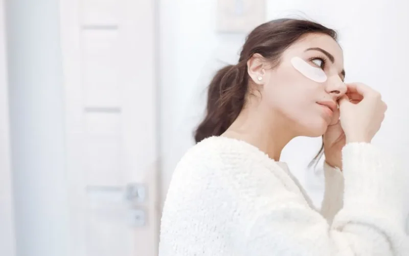 A woman in a cozy setting applying under eye masks as part of her skincare routine.