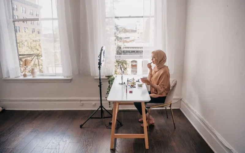 A Muslim woman applies makeup at a desk with natural light, creating content.