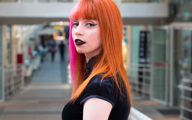 Stylish woman with orange and pink hair in an indoor architectural setting, showcasing bold fashion.