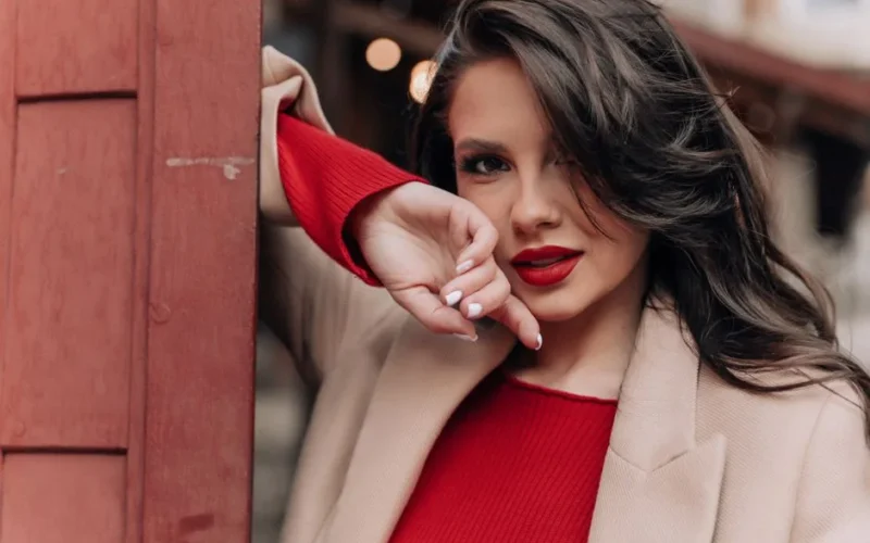 Portrait of an elegant woman with long brunette hair in a beige coat and red outfit, exuding fashion allure.