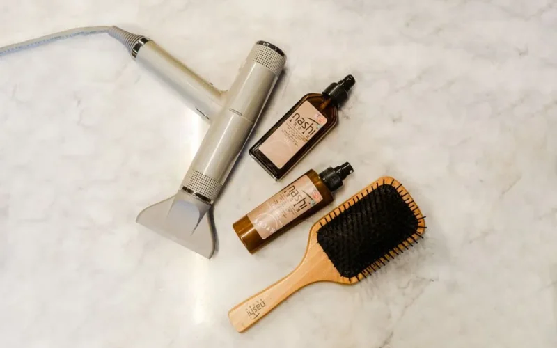 Flat lay of hairdryer, hairbrush, and styling products on a marble table.