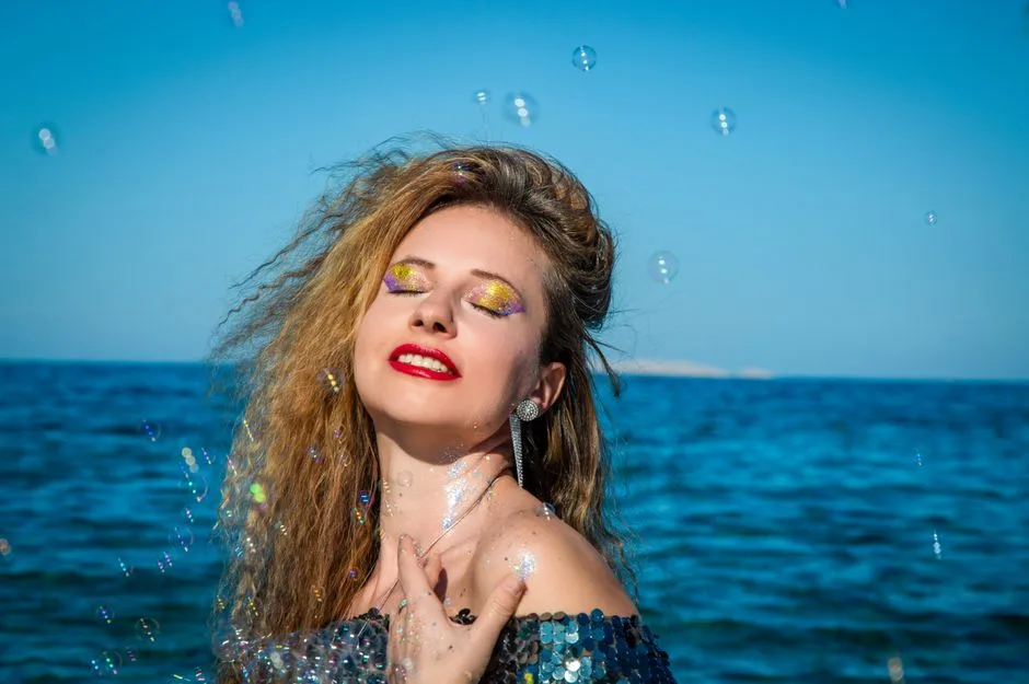 A vibrant portrait of a woman with glitter makeup and sequins enjoying a sunny day by the ocean.