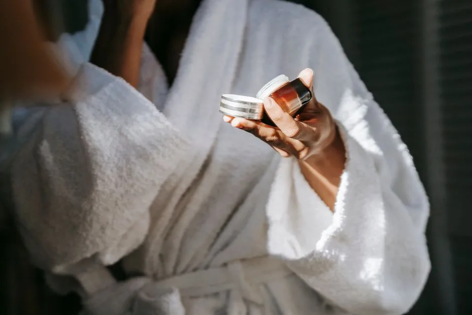 Crop anonymous ethnic female in bathrobe applying moisturizing cream on face while reflecting in mirror in house